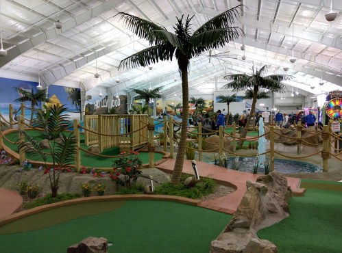 Indoor mini-golf course with palm trees, rocks, and colorful decorations, bustling with people enjoying the game.