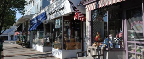Charming storefronts with American flags, featuring shops and benches along a sunny sidewalk.