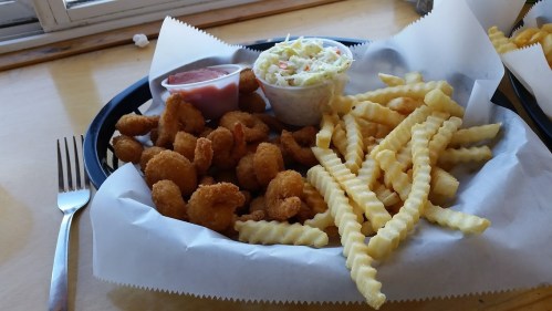 A plate of crispy fried shrimp, crinkle-cut fries, coleslaw, and dipping sauce on a paper-lined tray.