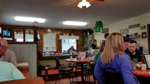 A cozy diner interior with patrons at tables, a counter, and festive decorations on the walls.