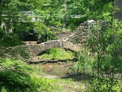 A stone arch bridge over a shallow stream, surrounded by lush greenery and trees.