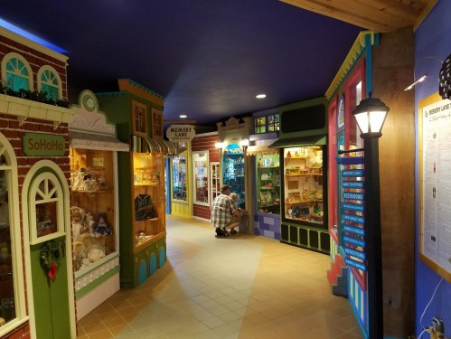 A colorful indoor display with shops and showcases, featuring a person examining items in a vibrant, whimsical setting.