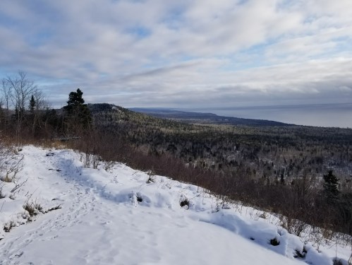 A snowy landscape with a view of a forested hillside and a distant body of water under a cloudy sky.
