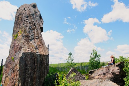 A person sits on a rock, gazing up at a large, towering rock formation under a blue sky with fluffy clouds.