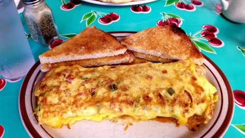 A plate with a fluffy omelette filled with vegetables and two slices of toasted bread on a colorful cherry-patterned table.