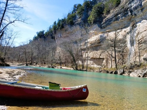 A red canoe rests on a sandy bank beside a clear, turquoise river, surrounded by rocky cliffs and trees.