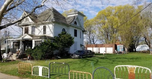 A large white Victorian house with a green lawn, surrounded by trees and old bed frames in the foreground.