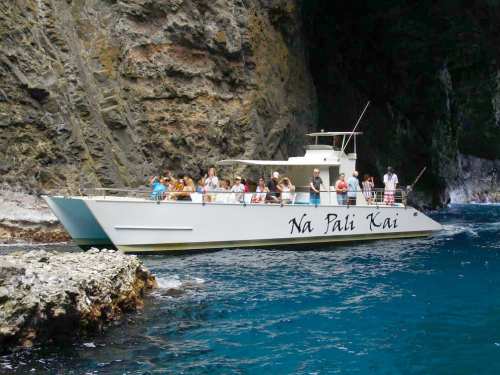 A white boat named "Na Pali Kai" with passengers, near rocky cliffs and clear blue water.