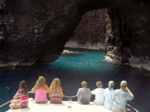 A group of children and adults sit on a boat, gazing at a rocky archway over turquoise water.