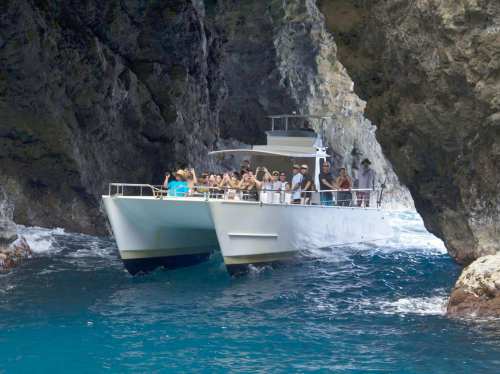 A large catamaran with passengers navigates through a rocky coastal passage, surrounded by clear blue water.