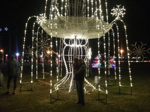 A couple stands under a large, illuminated fountain decorated with twinkling lights in a festive park setting.