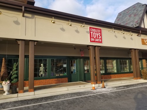 Exterior of a toy store with a sign reading "Coolest Toys on Earth," featuring large windows and decorative elements.