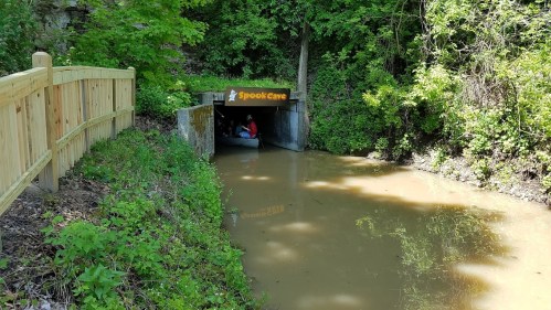 Entrance to Spook Cave surrounded by greenery, with a muddy waterway in front and a wooden fence nearby.
