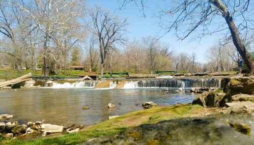 A serene river scene with a small waterfall, surrounded by trees and grassy banks under a clear blue sky.