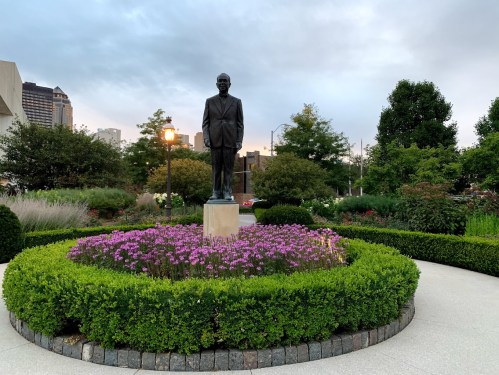 A statue of a man stands in a garden surrounded by colorful flowers and neatly trimmed hedges, with a city skyline in the background.