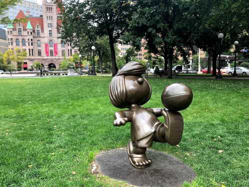 A bronze statue of a cartoon character kicking a ball in a park, surrounded by trees and buildings in the background.