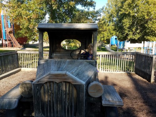 A child sits in the driver's seat of a wooden play truck at a park, surrounded by playground equipment and trees.