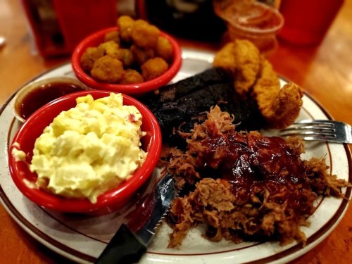 A plate of barbecue with pulled pork, fried chicken, potato salad, and fried okra, served with barbecue sauce.