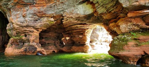 A natural rock formation with arches over green water, illuminated by sunlight filtering through the openings.