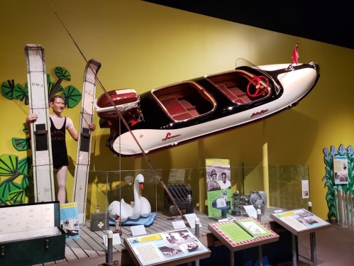 A vintage boat hangs above a display featuring water skis and historical photos in a colorful museum exhibit.