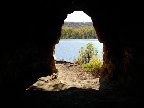 View from a cave opening overlooking a river and trees in the background on a sunny day.