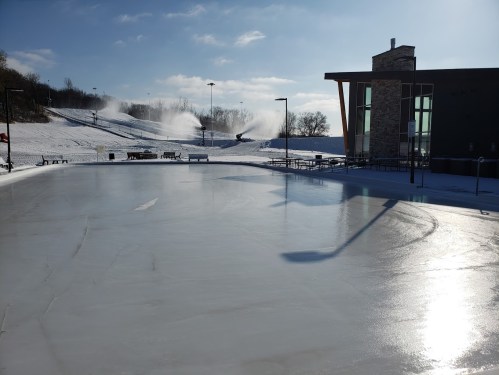 A sunny outdoor ice rink with snowmaking machines in the background and a clear blue sky.