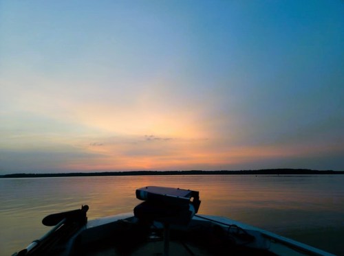 A serene sunset over calm waters, viewed from the front of a boat. Soft colors blend in the sky.