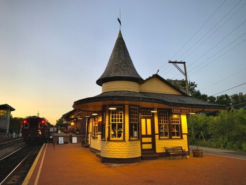 Historic train station at dusk, featuring a pointed roof and warm lights, with a train waiting on the tracks.