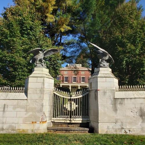 A grand entrance gate with eagle statues leads to a historic mansion surrounded by trees.