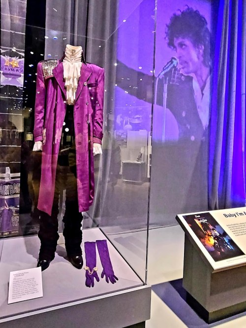 Purple outfit displayed in a glass case, with a backdrop of a performer on stage. A sign nearby provides information.