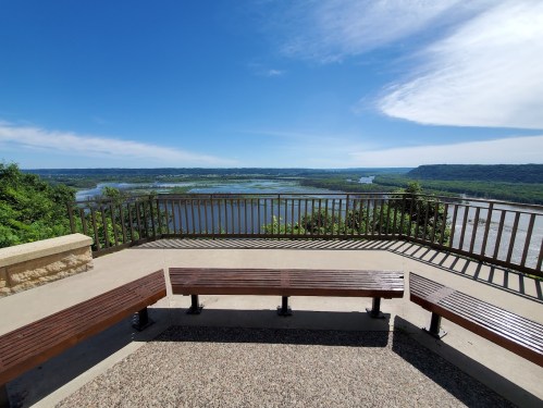 Scenic overlook with benches, featuring a river and lush hills under a clear blue sky.