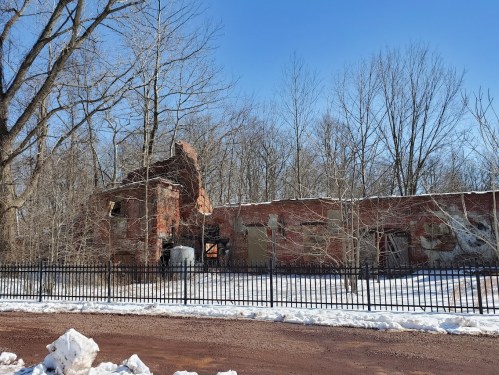 Ruins of a brick building surrounded by bare trees and snow, with a black fence in the foreground under a clear blue sky.