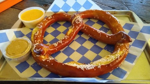A large, soft pretzel with coarse salt, served on a blue and white checkered paper, alongside mustard and cheese dips.