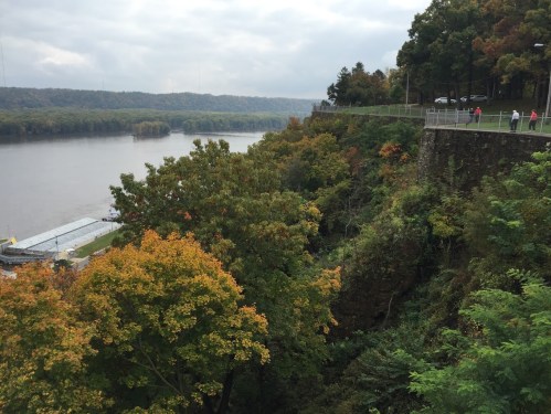 A scenic view of a river surrounded by autumn foliage and a rocky cliff, with a path and trees in the foreground.