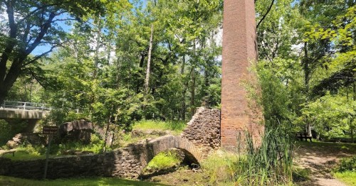 A stone bridge and a tall brick chimney surrounded by lush green trees in a serene outdoor setting.