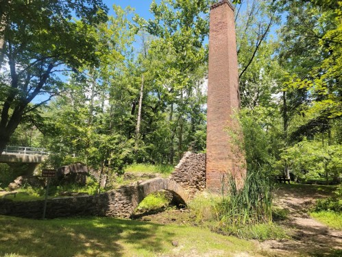 Ruins of a brick chimney and stone arch bridge surrounded by lush greenery and trees under a clear blue sky.