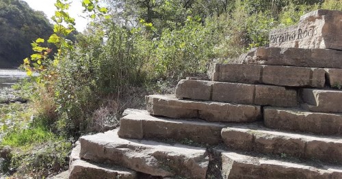 Stone steps lead to a natural area, surrounded by greenery and a river in the background.