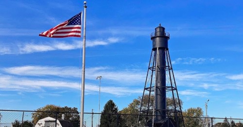 A tall black lighthouse beside an American flag, set against a clear blue sky with scattered clouds.