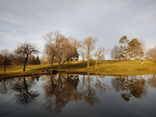 A serene pond reflecting trees and a house on a grassy hill under a cloudy sky.