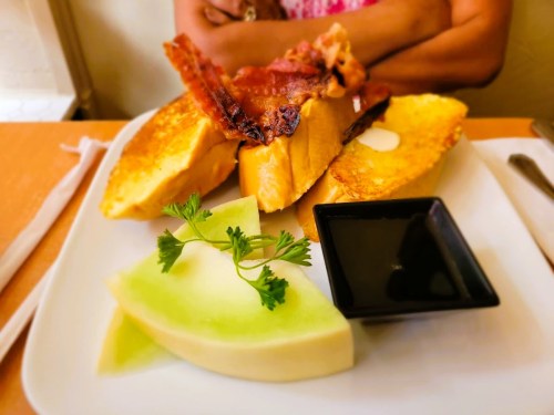 A plate of French toast topped with bacon, served with a slice of melon and a small dish of syrup.