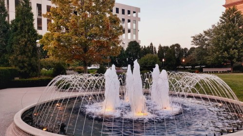 A fountain with water jets in a circular pool, surrounded by trees and buildings at sunset.