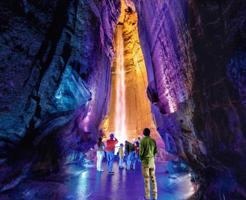 A group of people stands in a colorful cave, illuminated by vibrant lights, with a waterfall cascading in the background.