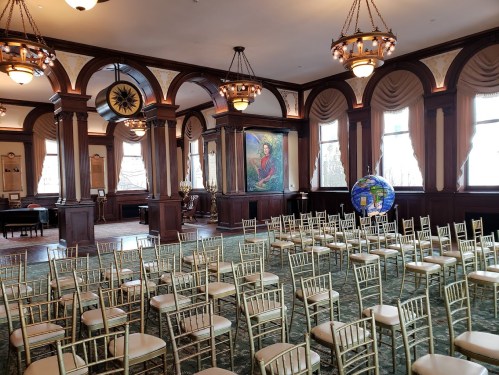 A spacious room with gold chairs arranged for an event, featuring large windows and a colorful mural on the wall.