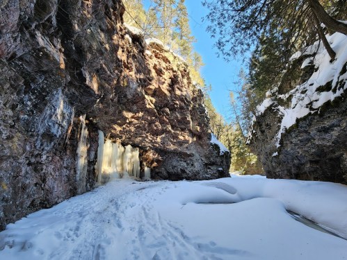 A snowy canyon with rocky walls and icicles hanging from the cliff, surrounded by trees under a clear blue sky.