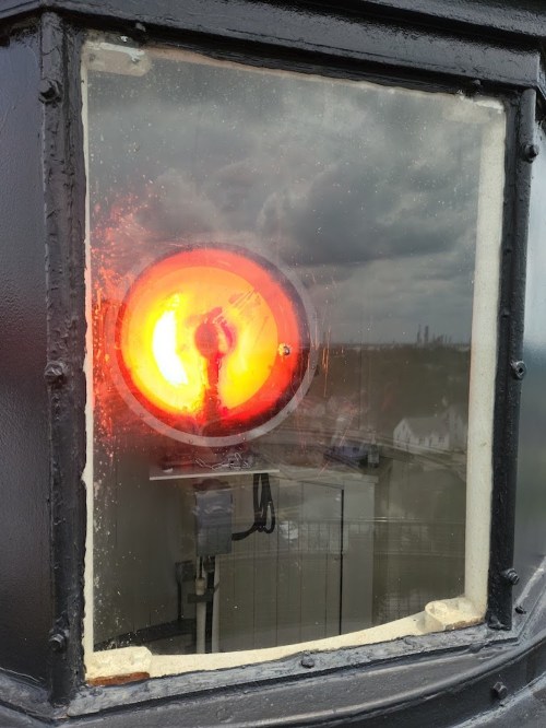 A close-up of a round orange light behind a glass panel, reflecting a cloudy sky and distant buildings.