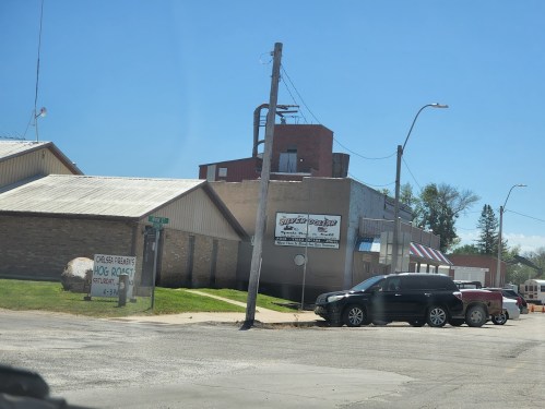 A street view of a small town with buildings, a sign for "Hog Rock," and parked vehicles on a sunny day.