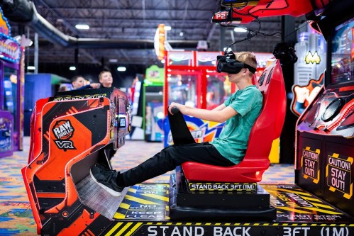 A person wearing a VR headset sits in a gaming chair at an arcade, focused on a virtual reality game.