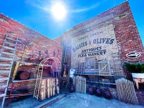 A rustic flea market entrance with vintage items, including ladders, pots, and wooden decor against a brick wall.