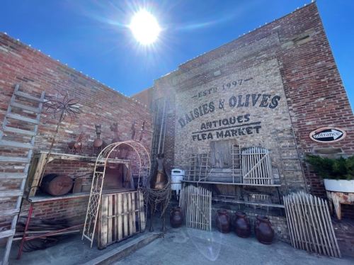 A rustic flea market with vintage items, ladders, and a sunlit brick wall featuring "Daisies & Olives" sign.