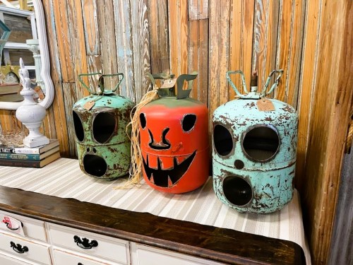 Three vintage gas canisters painted in bright colors, with one featuring a smiling pumpkin face, displayed on a wooden background.